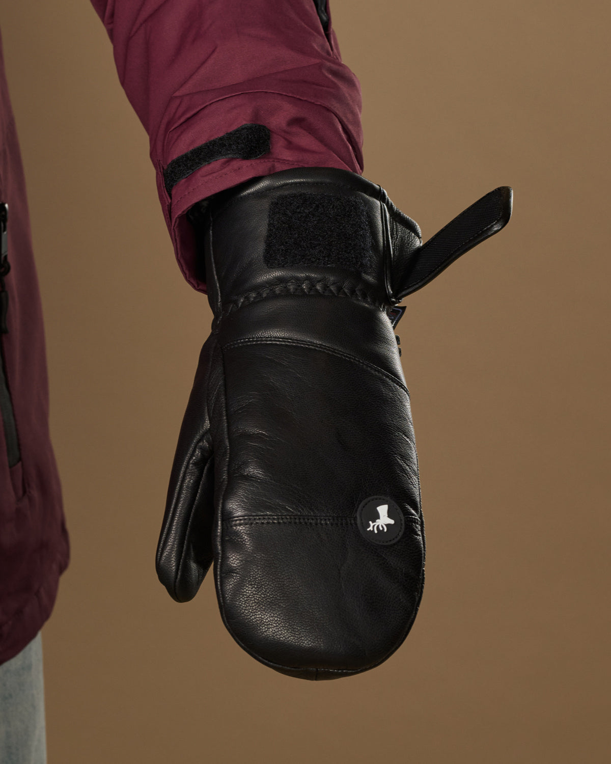 Black leather mitten close-up showing a wrist strap, stitched cuff, and logo patch.